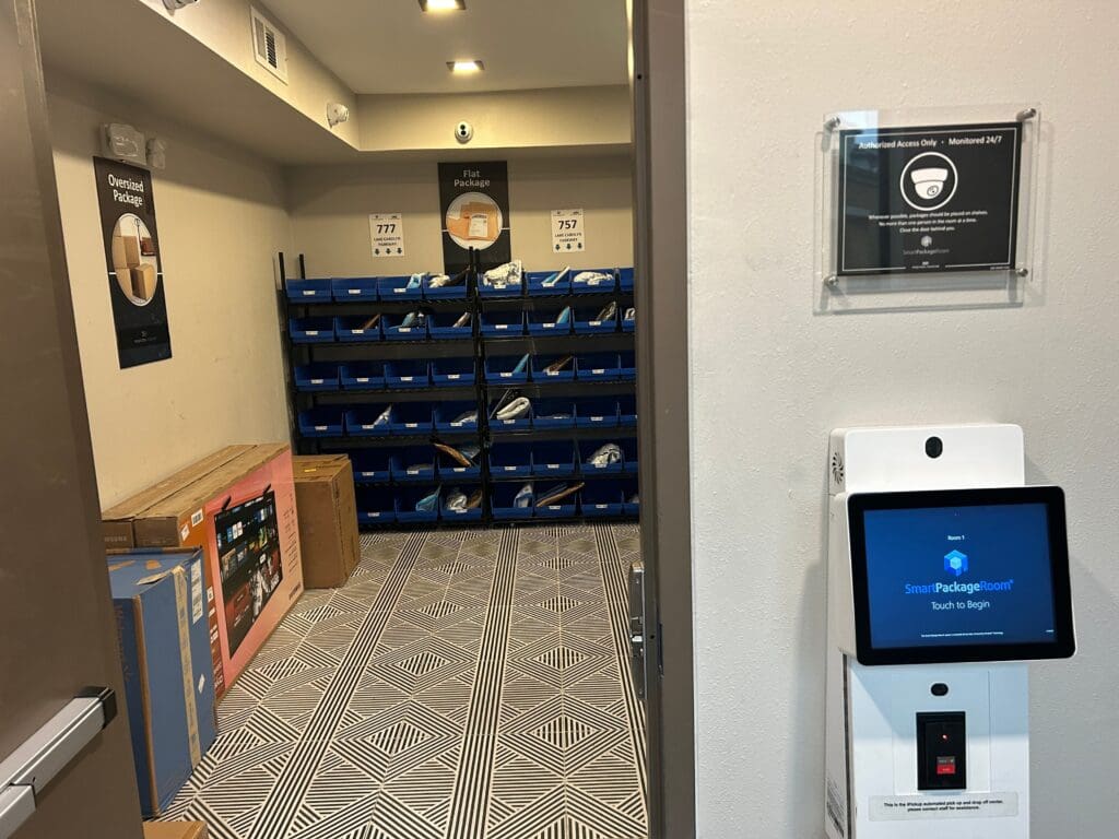 Smart Package Room with categorized areas for flat and oversized packages, 24/7 surveillance signage, and touchscreen access kiosk outside entry door. 