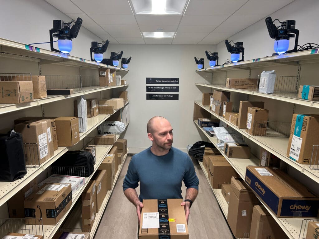 Resident retrieving a package inside a Smart Package Room equipped with overhead computer vision cameras and organized open shelving.