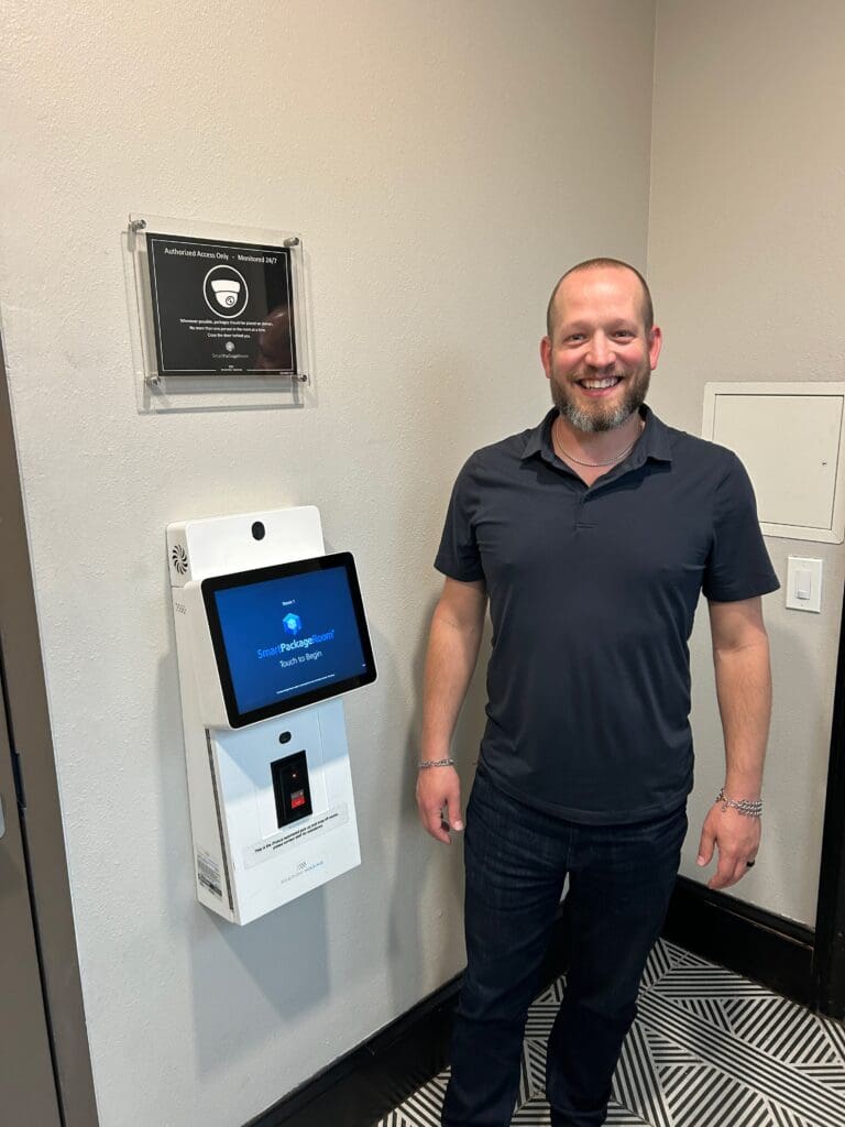 Smiling man standing next to a Smart Package Room touchscreen kiosk outside a monitored package room in a multifamily building.  
