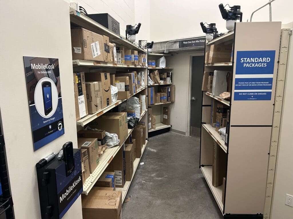 The Smart Package Room system with organized open shelving for standard packages, precision tracking technology, and a MobileKiosk device for couriers, showcasing secure, centralized package delivery in a multifamily building.