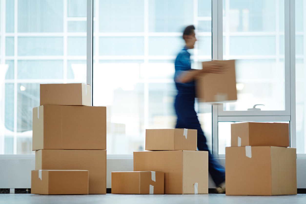 Delivery worker in blue uniform carrying a cardboard box past a large stack of packages in a bright lobby, illustrating the importance of choosing the right apartment package management system to avoid clutter.