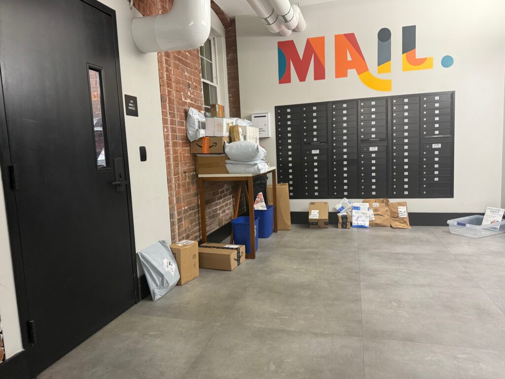 Cluttered apartment mailroom with packages overflowing onto the floor beside mailboxes under a MAIL mural
