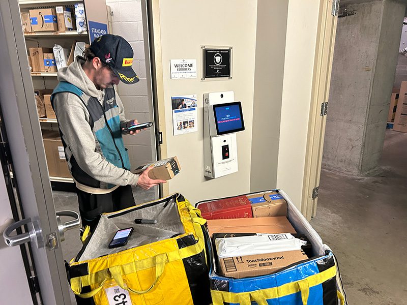 Amazon courier scanning a package at a Smart Package Room kiosk in an access-controlled, 24/7 monitored package room 